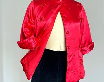 May include: A bright red satin jacket featuring a mandarin collar, long sleeves with folded cuffs, and a button-front closure. The fabric is decorated with a subtle, tonal embroidered floral pattern throughout the garment, displayed on a mannequin.