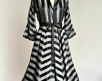 May include: A long-sleeved dress featuring a black and silver striped pattern. The bodice displays vertical stripes, while the skirt transitions into a chevron pattern. The garment includes a notched collar, three-quarter length sleeves, and a solid black fabric tie belt at the waist.