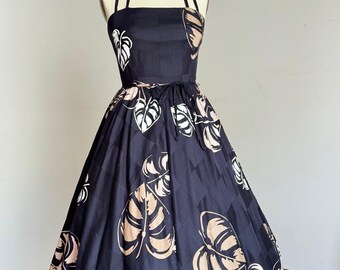 May include: A navy blue halter dress with a full skirt, featuring a repeating pattern of large leaves in white and tan. The dress has a fitted bodice and a tie at the waist. The dress is displayed on a mannequin.