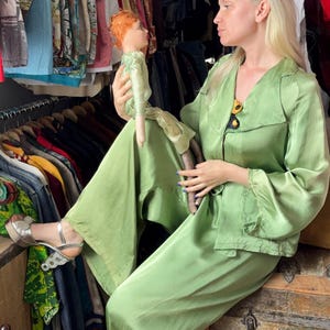 May include: A light green silk pajama set with a collared top and wide-leg pants. The model is holding a vintage doll with red hair and a green dress. The doll has a long, brown body. The model is wearing silver platform sandals.