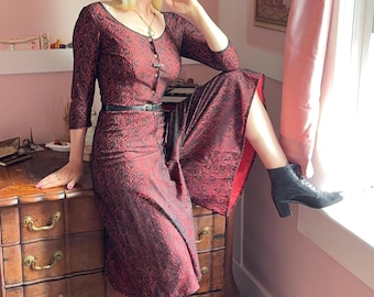 May include: A midi-length dress featuring a black and red floral lace overlay with a scoop neckline and three-quarter length sleeves. The dress includes a front button placket, a thin black belt at the waist, and a flared skirt with a red lining.
