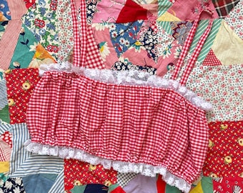 May include: A red and white gingham crop top featuring thin shoulder straps, a gathered elasticized bodice, and white lace trim detailing along the top and bottom edges of the garment, creating a textured and layered appearance.