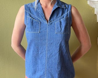 May include: A sleeveless blue denim shirt featuring a pointed collar, a V-neckline, and two square chest pockets. The garment is constructed with visible white stitching along the seams, pockets, and collar, providing a structured appearance.