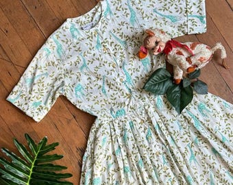 May include: A vintage white dress with a giraffe and leaf pattern in teal and olive green. The dress has short sleeves, a button-down front, and a pleated skirt. A vintage toy is on the dress.