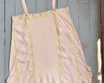 May include: A light pink camisole featuring vertical panels of cream-colored lace trim. The garment includes two thin shoulder straps and a ruffled lace hemline along the bottom edge, creating a layered appearance with a soft, smooth fabric finish.