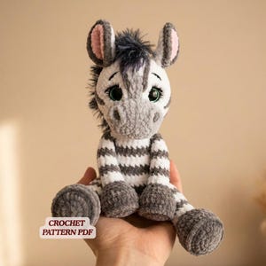 Könnte beinhalten: Ein handgehäkeltes Zebra-Plüschtier mit weiß-grau gestreiftem Körper, grauen Beinen und einem grau-weißen Kopf. Das Zebra hat große, ausdrucksstarke grüne Augen und rosa gefütterte Ohren. Der Text "CROCHET PATTERN PDF" ist sichtbar.