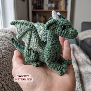 Könnte beinhalten: Ein handgefertigter grüner Häkchen-Drache mit weißen Hörnern und schwarzen Augen. Der Drache hat detaillierte Flügel und einen langen Schwanz. Der Text "CROCHET PATTERN PDF" ist sichtbar. Der Drache wird in einer Hand gehalten.