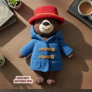May include: A handmade crochet bear wearing a blue coat with toggle closures and a red hat. The bear has a tan face and brown body. The image includes the text "CROCHET PATTERN PDF".
