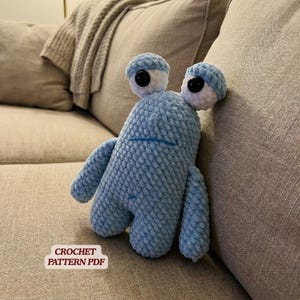 Op de afbeelding: Een lichtblauw gehaakt monsterknuffel met grote, ronde ogen en korte armen en benen. Het speelgoed is gemaakt van een zacht, getextureerd garen en heeft een gestikte mond. De tekst "CROCHET PATTERN PDF" staat in een witte doos.