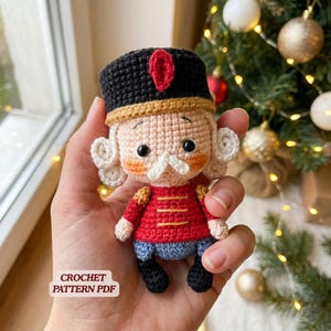 May include: A small crocheted nutcracker doll featuring a black hat with a red accent, a red jacket with yellow horizontal lines, blue trousers, and black boots. The doll has white curly hair, a white mustache, and rosy cheeks. Text reads CROCHET PATTERN PDF.