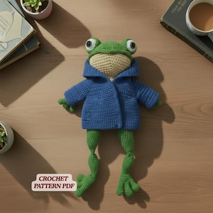 May include: A handmade crochet frog doll with a green head and legs, a tan face, and a blue jacket. The frog has large, round eyes and is posed on a wooden surface. The text "CROCHET PATTERN PDF" is visible.