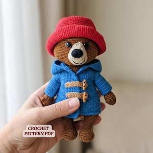 May include: A brown crocheted bear toy wearing a bright red hat and a blue hooded duffle coat with two tan toggle closures. The bear has black eyes and a black nose. A small white rectangular label with the text CROCHET PATTERN PDF is visible.