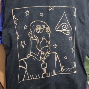 Peut inclure: Un t-shirt noir à manches courtes avec un dessin au trait clair représentant une personne chauve au nez proéminent portant une chemise à col. Le motif est encadré par un carré avec des étoiles et un symbole triangulaire en haut à droite.