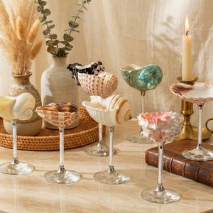 May include: A display of seashell goblets with crystal stems. The shells are in various colours and shapes, including white, brown, and iridescent green. A lit candle sits in a brass holder, and a woven tray is in the background.