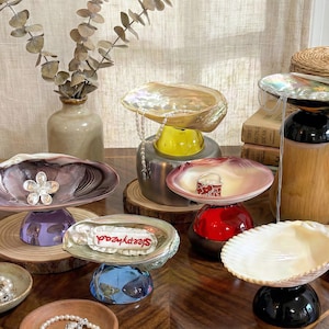 Peut inclure: Une collection de plats et de bols décoratifs en forme de coquillage de différentes couleurs, dont irisé, violet et blanc. Certains coquillages sont montés sur des socles colorés et certains ont des éléments décoratifs comme des perles et le mot "Sleepyhead".