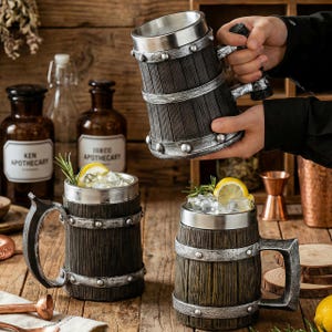 Könnte beinhalten: Bierkrüge in Holzoptik mit silberfarbenen Akzenten. Die Krüge haben ein Fassdesign mit Metallbändern und Nieten. Einer wird gehalten, während zwei weitere auf einer Holzoberfläche stehen, gefüllt mit Eis, Zitronenscheiben und Kräutern.