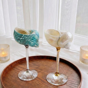 Puede incluir: Dos copas de vino decorativas con forma de concha. Una es turquesa y blanca, la otra blanca nacarada. Tienen tallos de cristal y están sobre una bandeja de madera. Hay velas cerca, creando una estética costera y elegante.