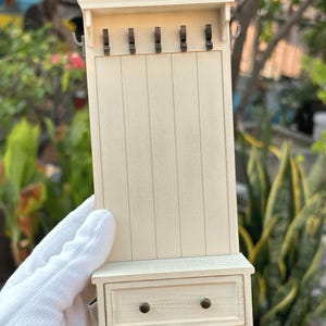 May include: A miniature off-white hall tree with a drawer and five hooks. The small furniture piece has a rectangular design with vertical paneling and a small shelf above the drawer. The hooks are dark metal.