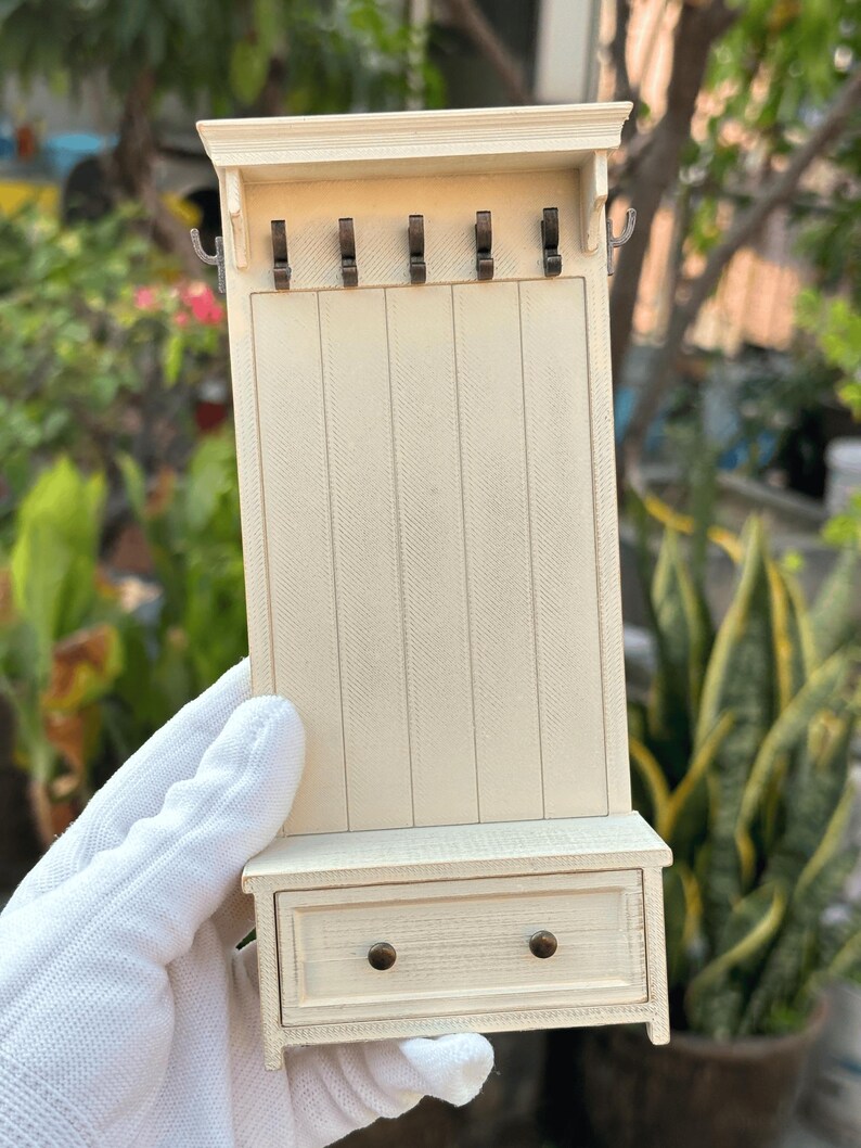 May include: A miniature off-white hall tree with a drawer and five hooks. The small furniture piece has a rectangular design with vertical paneling and a small shelf above the drawer. The hooks are dark metal.