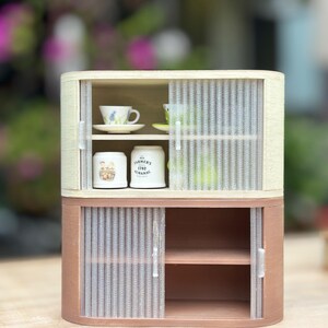 May include: A miniature two-tiered cabinet with sliding doors. The top section has a light wood frame and houses a teacup and saucer. The bottom section is a darker wood tone with shelves. The cabinet contains two small white jars with text.