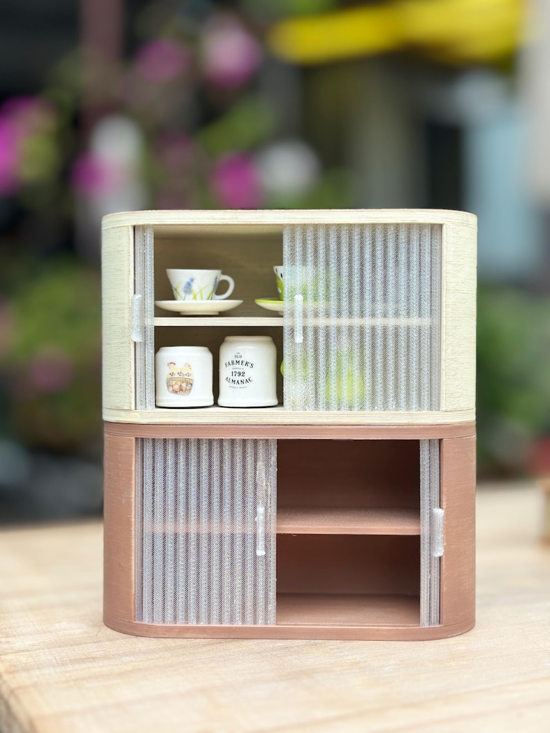 May include: A miniature two-tiered cabinet with sliding doors. The top section has a light wood frame and houses a teacup and saucer. The bottom section is a darker wood tone with shelves. The cabinet contains two small white jars with text.