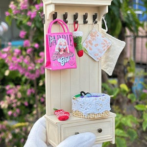 May include: Miniature off-white coat rack with hooks, a drawer, and a shelf. A pink Barbie Girl tote bag, a green and red stocking, and two fabric squares hang from the hooks. A small basket and a red knitted item sit on the shelf.