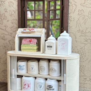 May include: Miniature kitchen scene with a wooden cabinet, jars, and a window. The cabinet has a sliding door and holds various jars labeled "Sugar," "Fresh Coffee," and "The Cat Bakery." A small display case holds miniature baked goods.