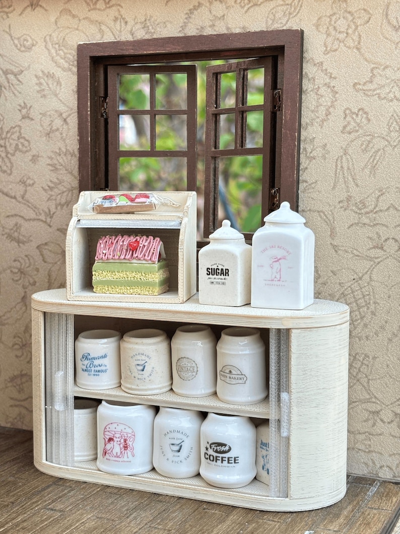 May include: Miniature kitchen scene with a wooden cabinet, jars, and a window. The cabinet has a sliding door and holds various jars labeled "Sugar," "Fresh Coffee," and "The Cat Bakery." A small display case holds miniature baked goods.