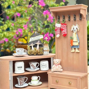 May include: A miniature wooden display featuring a small cabinet with teacups, sugar, and coffee containers. A matching coat rack holds decorative stockings and a bunny figure. A small teddy bear sits on a bench below.