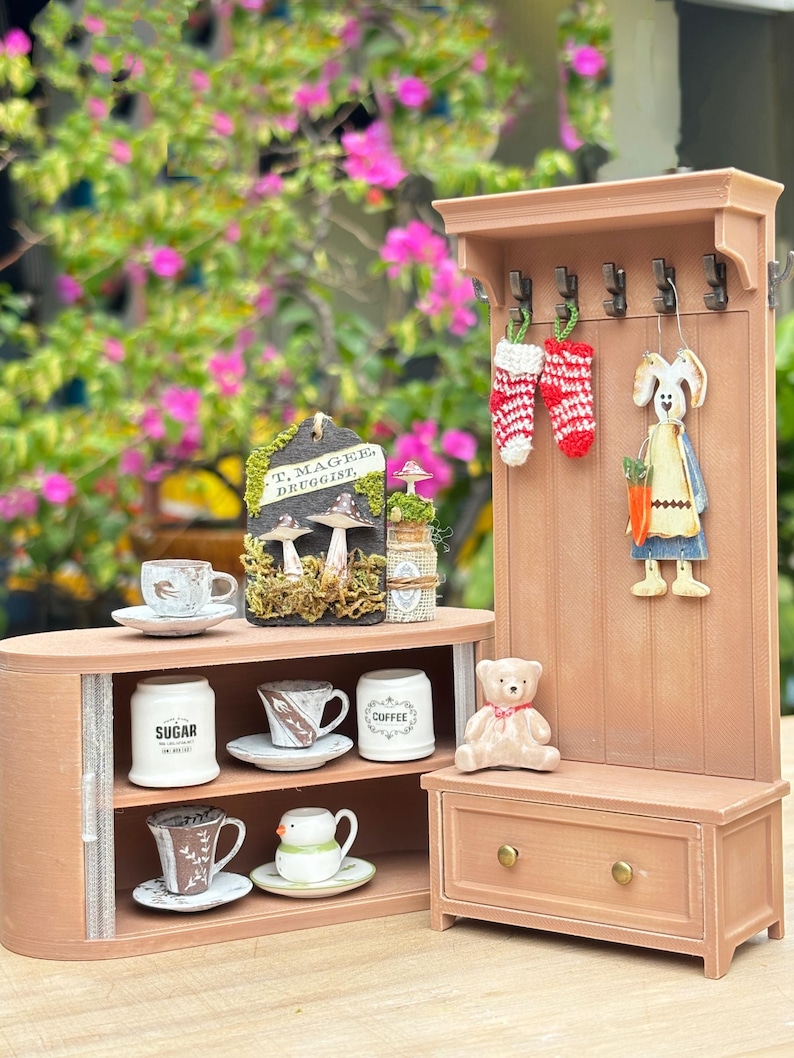 May include: A miniature wooden display featuring a small cabinet with teacups, sugar, and coffee containers. A matching coat rack holds decorative stockings and a bunny figure. A small teddy bear sits on a bench below.