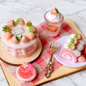 Puede incluir: Miniaturas de postres sobre una bandeja de madera. Un pastel rosa con fresas, un vaso con crema batida, una gelatina rosa y un brazo de gitano rosa. Un tenedor dorado descansa sobre una servilleta a cuadros rojos y blancos.