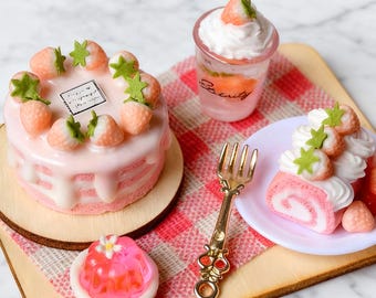 May include: Miniature desserts on a wooden tray. A pink cake with strawberry toppings, a glass with whipped cream, a pink jelly, and a pink roll cake with strawberries. A gold fork rests on a red and white checkered napkin.