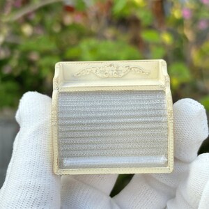 May include: A small, rectangular, off-white antique box with a decorative top and a clear, ribbed front. The box is held in a gloved hand against a blurred green and pink background.
