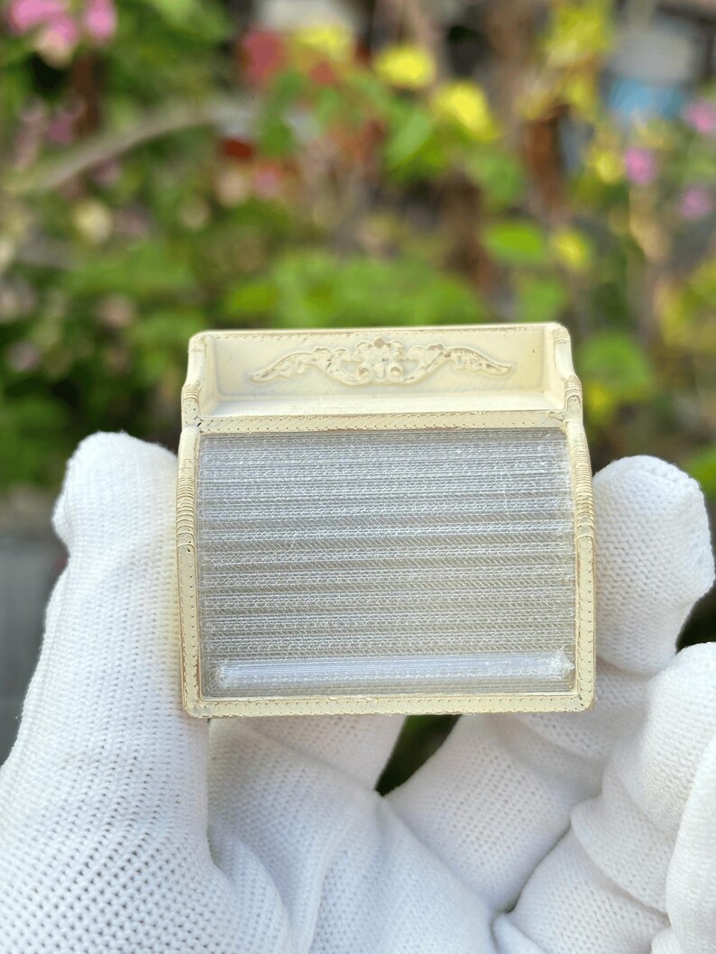 May include: A small, rectangular, off-white antique box with a decorative top and a clear, ribbed front. The box is held in a gloved hand against a blurred green and pink background.