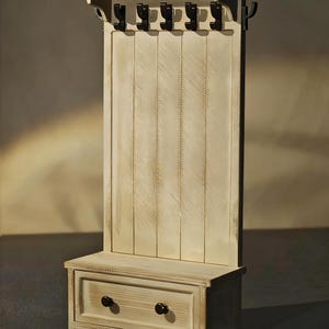 May include: A cream-colored wooden hall tree with a bench, drawer, and coat hooks. The hall tree has a distressed finish, with six black metal hooks and a drawer with two dark knobs. The bench is at the base of the hall tree.