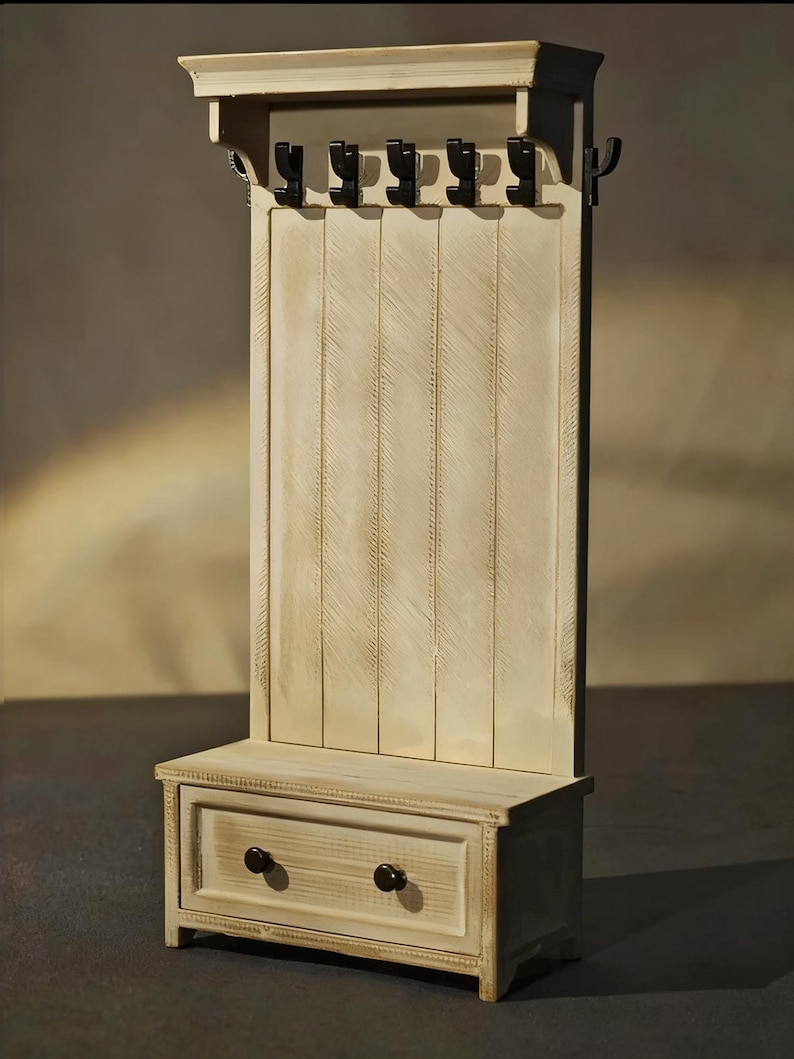 May include: A cream-colored wooden hall tree with a bench, drawer, and coat hooks. The hall tree has a distressed finish, with six black metal hooks and a drawer with two dark knobs. The bench is at the base of the hall tree.