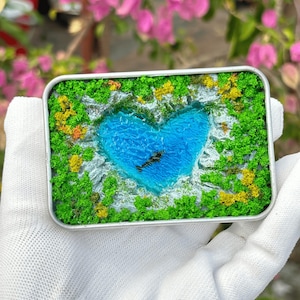 May include: A miniature landscape scene in a rectangular metal frame. The scene features a heart-shaped blue lake surrounded by green and yellow foliage. The frame is held in a gloved hand, with pink flowers in the background.