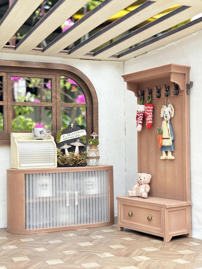 May include: A miniature dollhouse interior scene featuring a wooden cabinet, a coat rack with decorative items, and a small bench. The scene includes a teddy bear, a bunny figurine, and a sign that reads "T. Magee, Druggist."