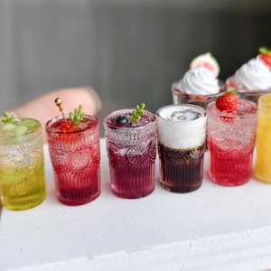 May include: A collection of six miniature decorative glasses filled with colorful liquid beverages. The glasses feature textured patterns and are topped with small garnishes including berries, whipped cream, and green sprigs.