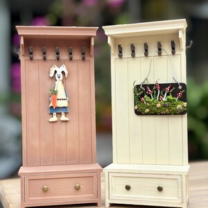 May include: Two decorative miniature coat racks. One is painted a light pink with a bunny figure holding a carrot. The other is cream-colored with a small sign featuring mushrooms. Both have drawers and hooks.