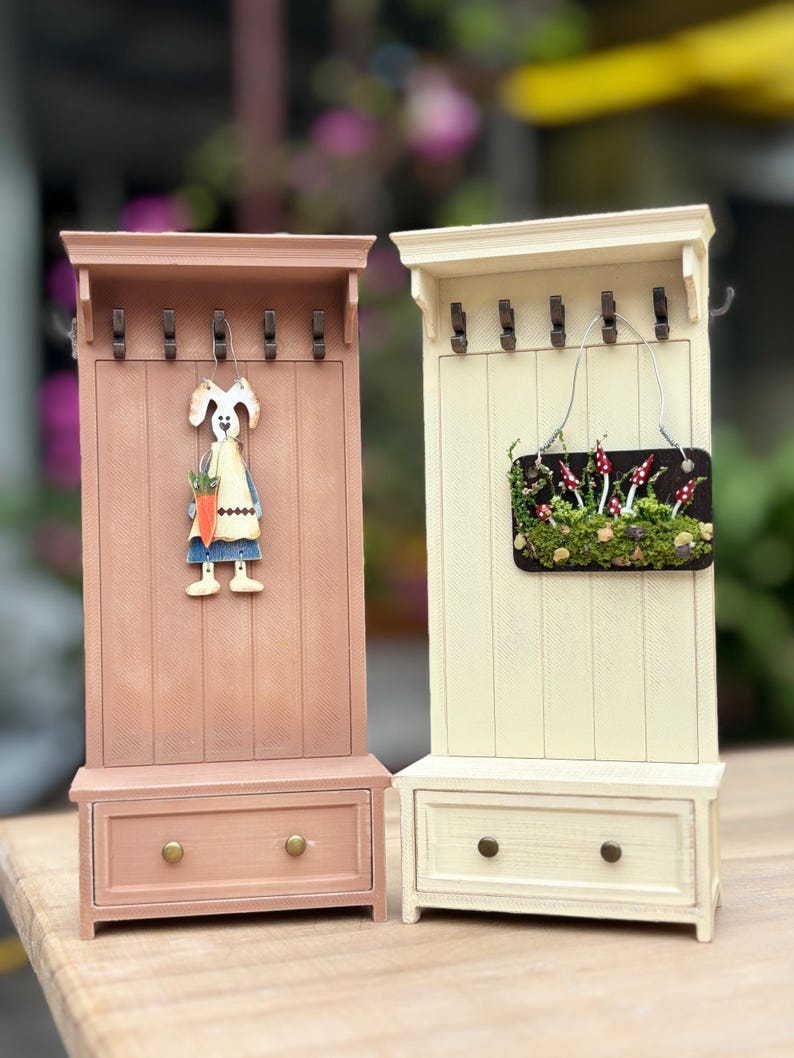 May include: Two decorative miniature coat racks. One is painted a light pink with a bunny figure holding a carrot. The other is cream-colored with a small sign featuring mushrooms. Both have drawers and hooks.