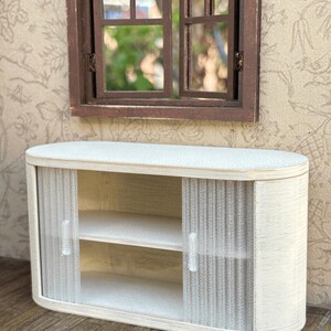 May include: A miniature white cabinet with sliding doors and two shelves. The cabinet has a rounded shape and is made of wood. The doors are made of a textured, semi-transparent material. A brown window is above the cabinet.
