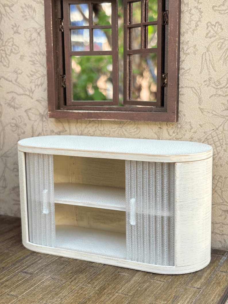 May include: A miniature white cabinet with sliding doors and two shelves. The cabinet has a rounded shape and is made of wood. The doors are made of a textured, semi-transparent material. A brown window is above the cabinet.