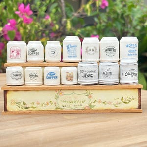 Puede incluir: Una colección de pequeños tarros de cerámica blanca con varias etiquetas, incluyendo "Coffee" y "Showcase". Los tarros están dispuestos en una estantería de madera con detalles florales y la inscripción "hand made".