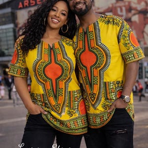 May include: Two matching short-sleeved shirts featuring a vibrant yellow base with intricate orange, green, and black geometric patterns. The shirts have a central vertical design, a rounded neckline, and a straight hemline, paired with black jeans.
