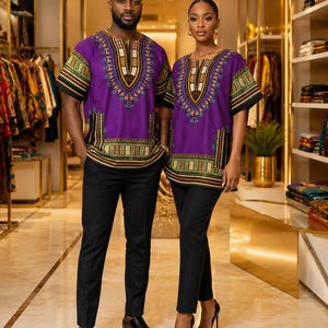 May include: Two individuals wearing matching purple dashiki shirts featuring intricate geometric patterns in yellow, green, and black around the neckline, sleeves, and hem. Both shirts are paired with solid black trousers, creating a coordinated look.