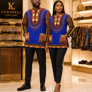 May include: A vibrant blue long-sleeve tunic featuring a colourful, intricate geometric pattern around the neckline, cuffs, and hem, with two front pockets. The garment is paired with solid black trousers and black dress shoes.