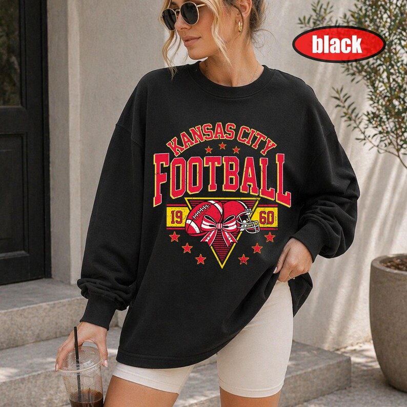 May include: A black long-sleeve sweatshirt featuring a graphic print with the text KANSAS CITY FOOTBALL in yellow and red. The design includes a football, a helmet, a bow, and the numbers 19 and 60, all centred within a yellow triangle.