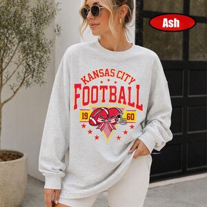 May include: An ash grey long-sleeve sweatshirt featuring a red and yellow graphic print that reads Kansas City Football with the numbers 19 and 60. The design includes a football helmet with a red bow and several small red stars.