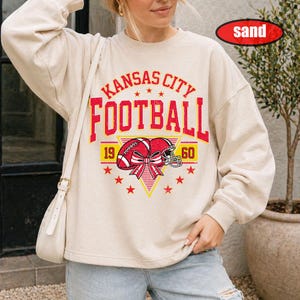 May include: A sand-coloured crewneck sweatshirt featuring a distressed red and yellow graphic print. The design includes the text KANSAS CITY FOOTBALL, the numbers 19 and 60, a football, a helmet, a decorative bow, and several small star icons.